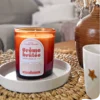 Bougie parfumée Crème Brûlée en verre ambré posée sur un plateau blanc, ambiance intérieure cosy avec vase décoratif et tasse sur une table en fibres naturelles.