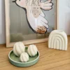 Ambiance déco avec des bougies spirales artisanales et un brûle-parfum en forme d’arc-en-ciel sur une étagère en bois. Mise en scène minimaliste avec une affiche 'Peace on Earth' et un plateau vert pastel.