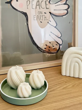 Ambiance déco avec des bougies spirales artisanales et un brûle-parfum en forme d’arc-en-ciel sur une étagère en bois. Mise en scène minimaliste avec une affiche 'Peace on Earth' et un plateau vert pastel.
