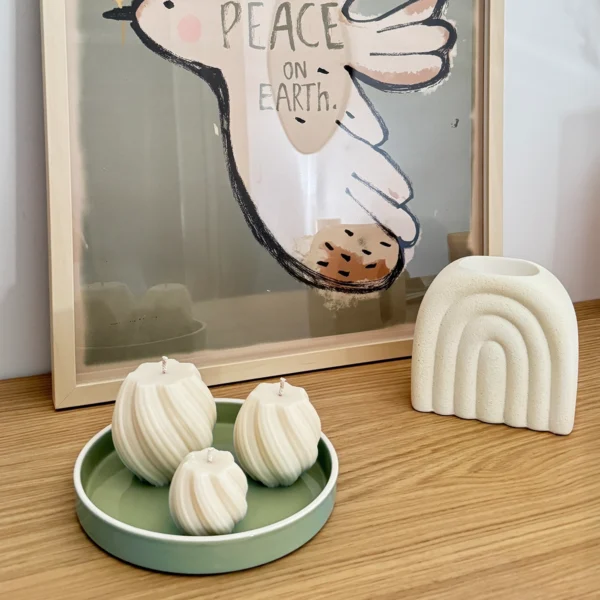 Ambiance déco avec des bougies spirales artisanales et un brûle-parfum en forme d’arc-en-ciel sur une étagère en bois. Mise en scène minimaliste avec une affiche 'Peace on Earth' et un plateau vert pastel.