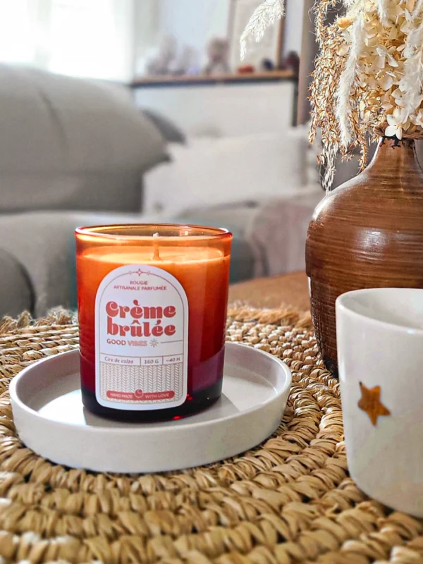 Bougie parfumée Crème Brûlée en verre ambré posée sur un plateau blanc, ambiance intérieure cosy avec vase décoratif et tasse sur une table en fibres naturelles.