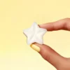 Fondant parfumé en forme d’étoile, parfum crème brûlée, tenu entre deux doigts avec vernis jaune, sur fond jaune pastel