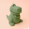 Bougie verte en forme de dinosaure T-Rex rigolo, posée sur fond violet pastel.