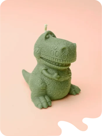 Bougie verte en forme de dinosaure T-Rex rigolo, posée sur fond violet pastel.