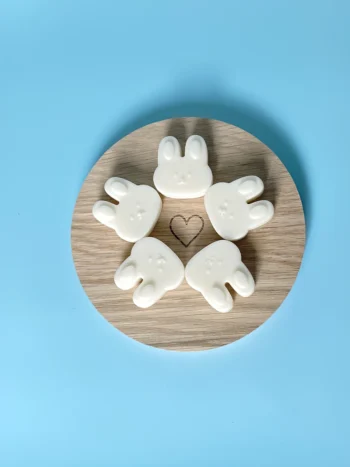 Fondants parfumés en forme de tête de lapin, parfum Floraison, disposés en cercle sur un support en bois rond avec un cœur gravé, sur fond bleu clair – Ambiance printanière et douce.