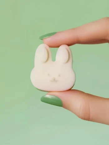 Fondant Floraison – Parfum Printanier (Lapin) Fondant parfumé en forme de tête de lapin blanc, tenu entre deux doigts avec vernis vert pastel, sur fond vert, parfum printanier