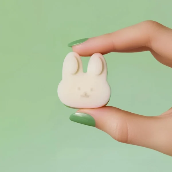 Fondant parfumé en forme de tête de lapin blanc, tenu entre deux doigts avec vernis vert pastel, sur fond vert, parfum printanier