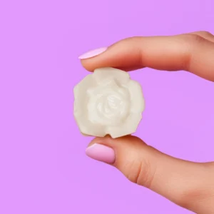 Fondant parfumé en forme de rose blanche, tenu entre deux doigts avec vernis rose pastel, sur fond violet, Rosebud