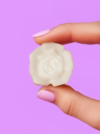 Fondant parfumé en forme de rose blanche, tenu entre deux doigts avec vernis rose pastel, sur fond violet, Rosebud
