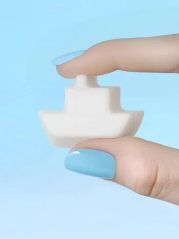Fondant parfumé Flow en forme de bateau en cire de colza Fondant parfumé en forme de bateau tenu entre deux doigts vernis bleu, sur fond bleu pastel.