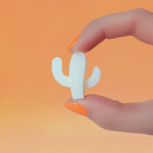 Fondant parfumé en forme de cactus blanc, tenu entre deux doigts vernis orange, sur fond orange dégradé.