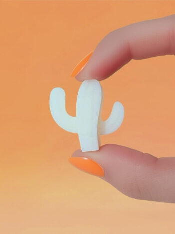 Fondant parfumé en forme de cactus blanc, tenu entre deux doigts vernis orange, sur fond orange dégradé.