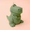 Bougie verte en forme de dinosaure T-Rex rigolo, posée sur fond violet pastel.