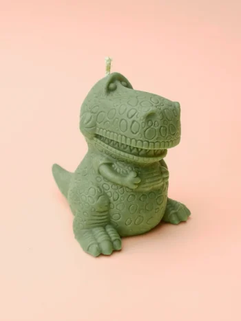 Bougie verte en forme de dinosaure T-Rex rigolo, posée sur fond violet pastel.
