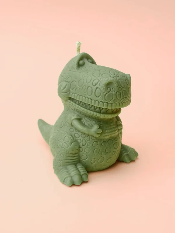 Bougie verte en forme de dinosaure T-Rex rigolo, posée sur fond violet pastel.