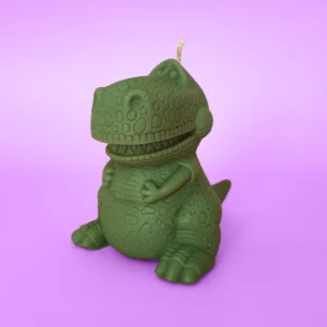 Bougie verte en forme de dinosaure T-Rex rigolo, posée sur fond violet pastel.