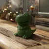 Bougie verte en forme de dinosaure T-Rex rigolo, posée sur une table en bois dans une ambiance cosy.