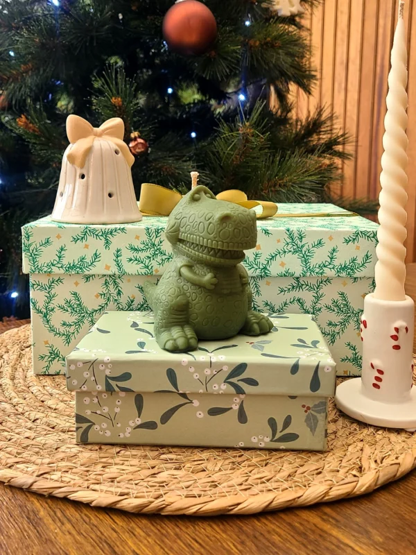 Bougie décorative verte en forme de dinosaure Roooaar, posée sur des boîtes cadeaux devant un sapin de Noël.